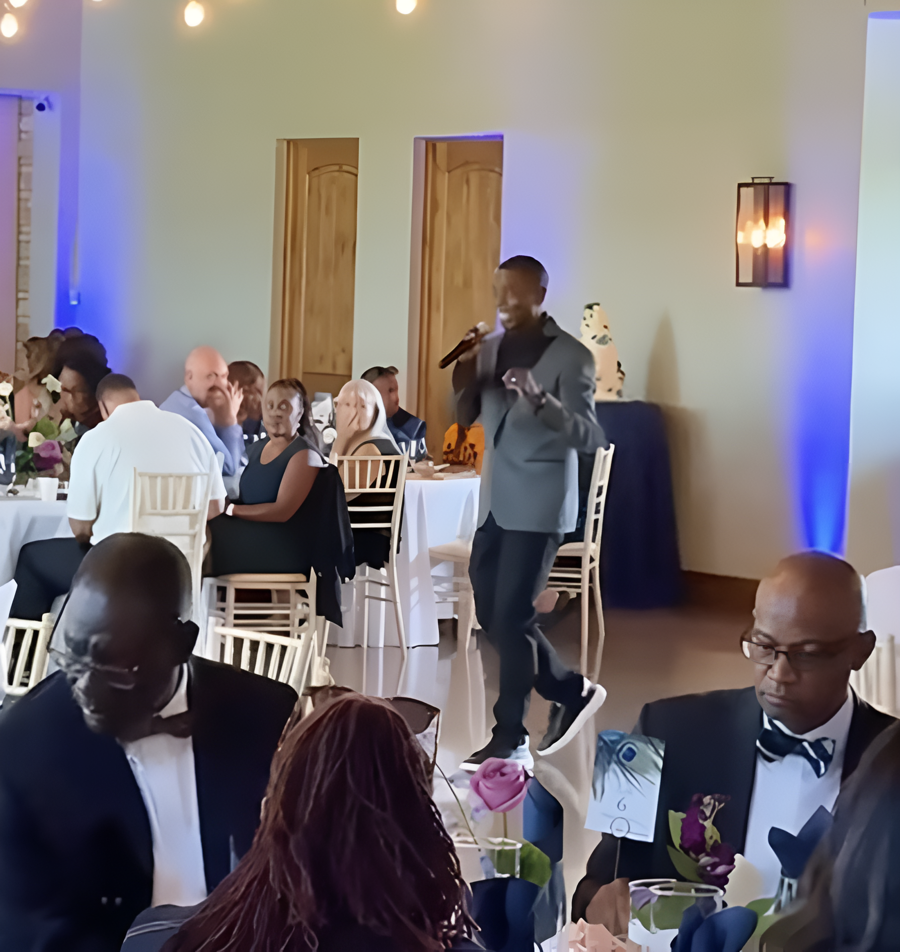 A man speaks into a microphone at an event, surrounded by seated guests at tables. The room has warm lighting and a neutral color scheme.