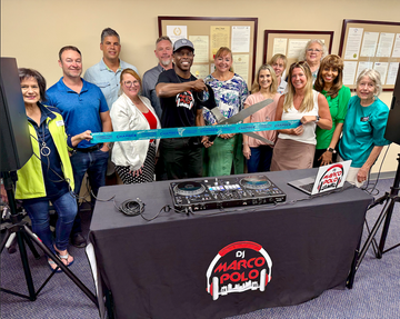 A group of people at a DJ setup, smiling while holding giant scissors for a ribbon-cutting ceremony.