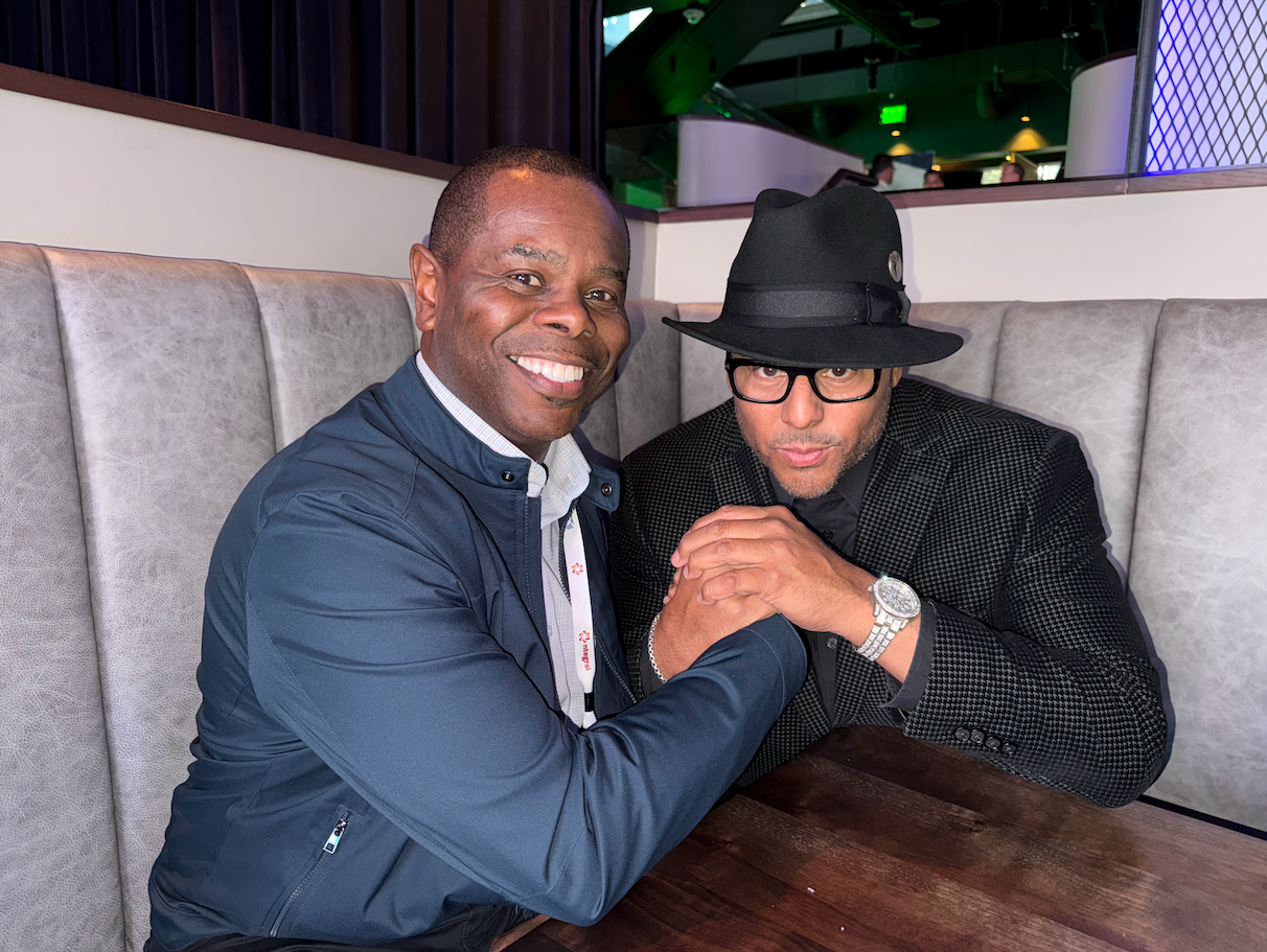 Two men smiling at a table. One wears a blue jacket, the other a hat and black patterned jacket. They're indoors with a cushioned booth.