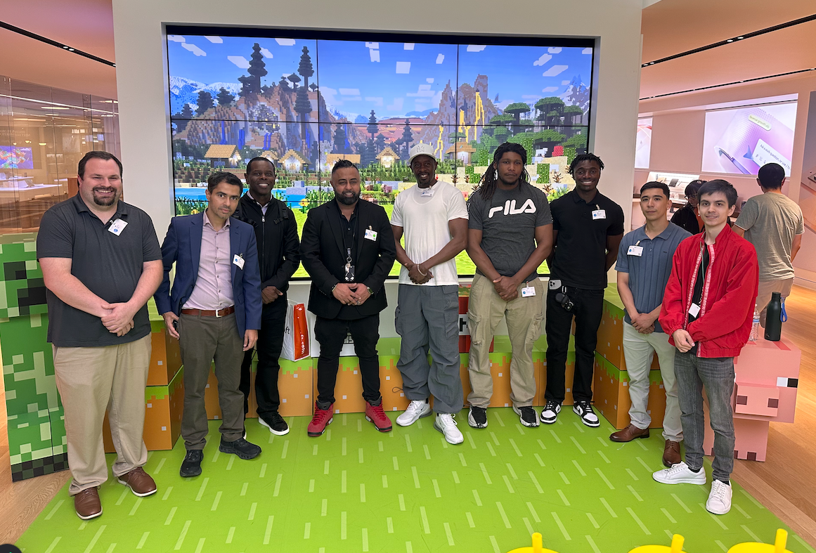 Group of people posing for a photo in front of a large Minecraft screen. They stand on green turf with Minecraft-themed decorations in a brightly lit space.