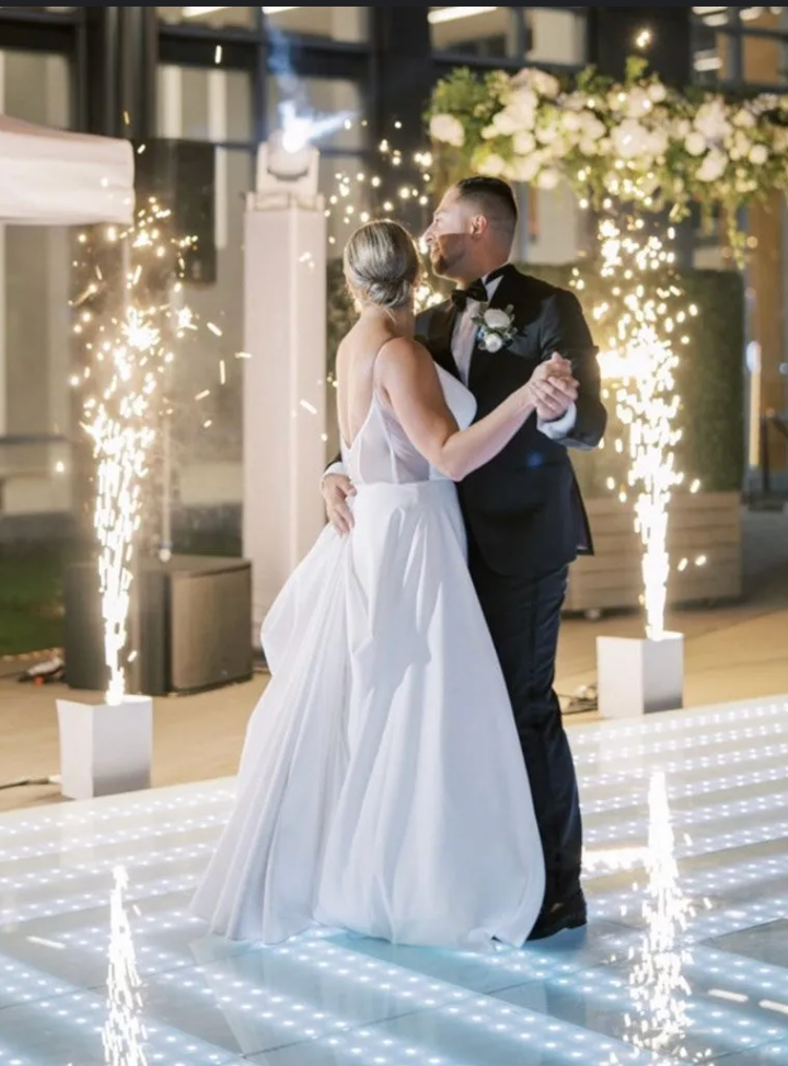 A newlywed couple dances on a sparkling white dance floor, with celebratory fireworks erupting around them. They are outdoors at night.