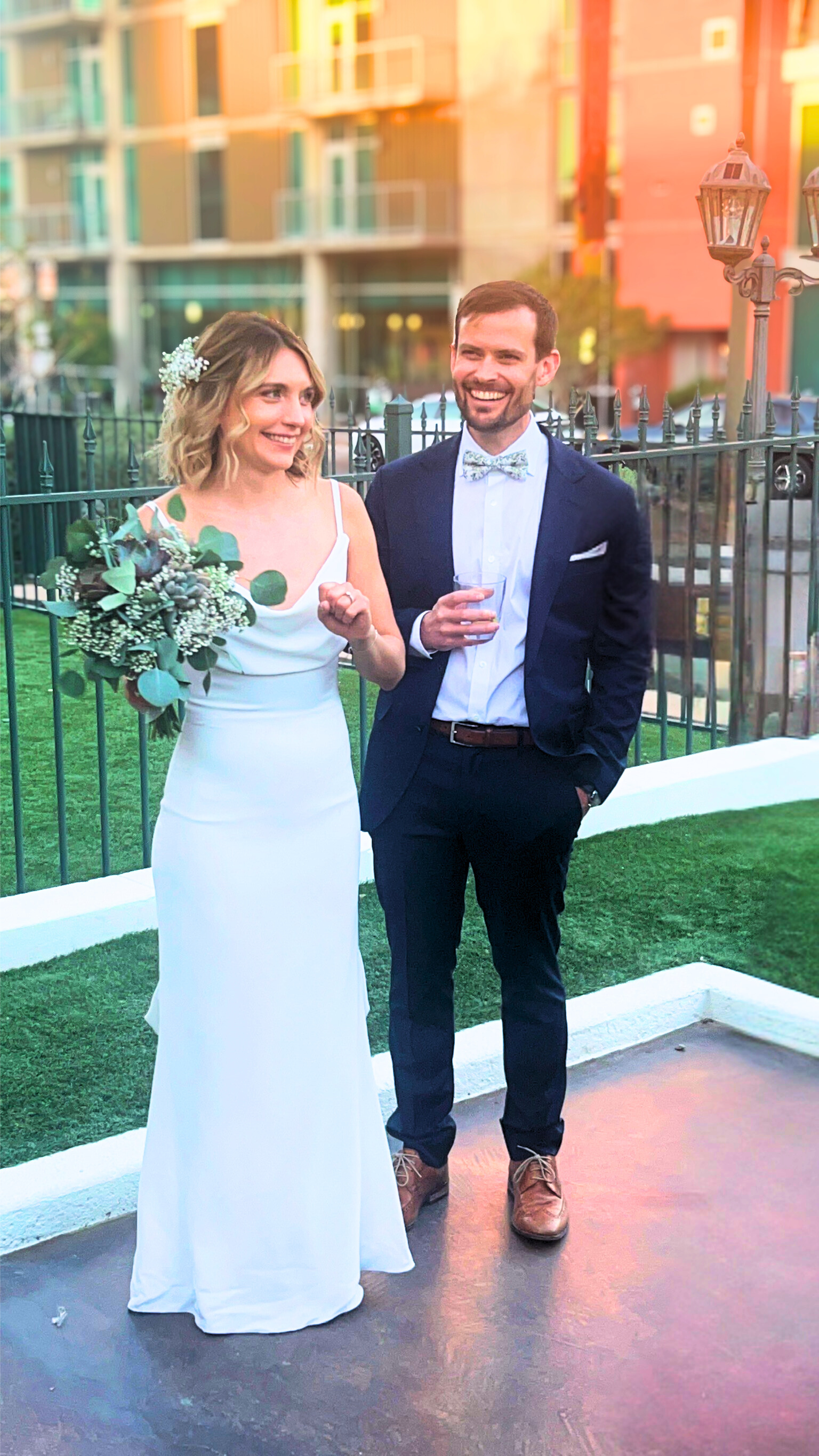 Bride and groom smiling at each other. The woman in a white dress, the man in a suit. They stand outside, by a fence.