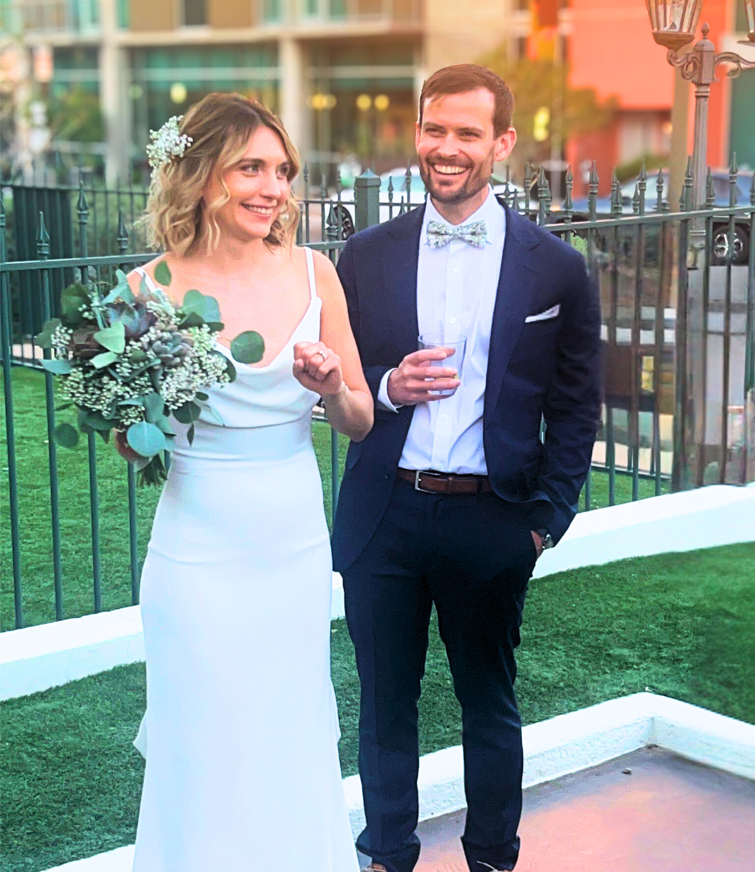 A newly married couple smiles outdoors. The bride wears a white dress, and the groom a navy suit and bowtie.