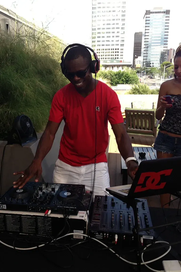 A DJ wearing sunglasses and a red shirt smiles while mixing music outdoors. He's surrounded by equipment, with a cityscape in the background.