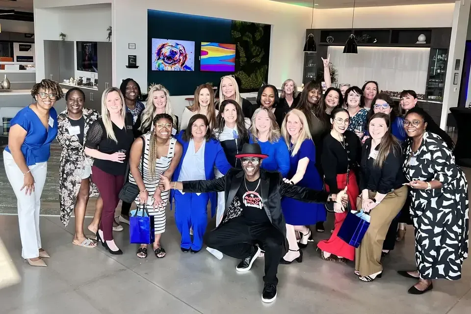 A large group of diverse women and one man pose inside a modern building. The man kneels in the front wearing a hat and graphic tee.