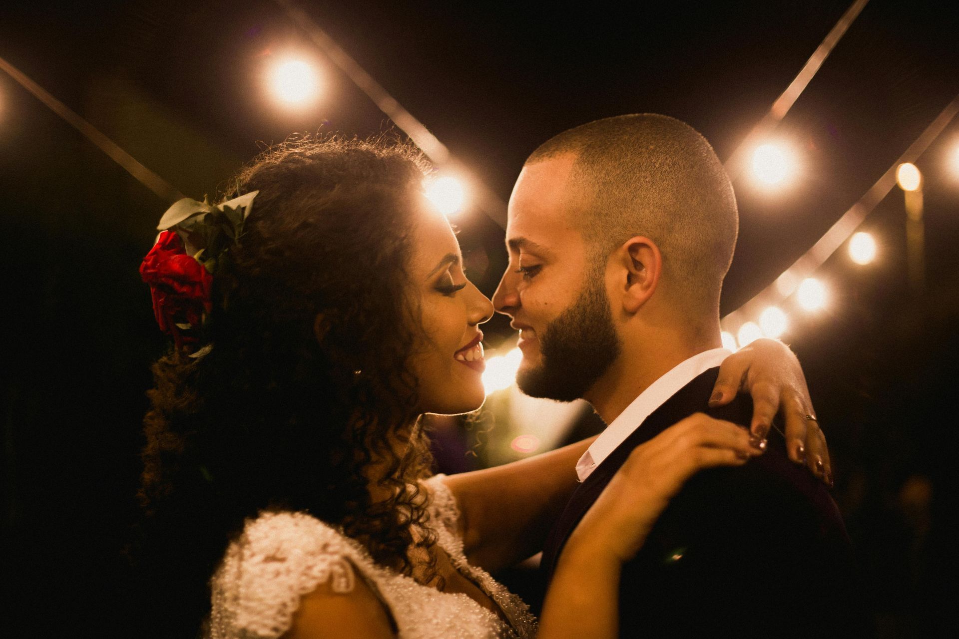 Bride and groom embrace and gaze at each other intimately under string lights. Woman wears a white lace dress and has a red rose in her hair.