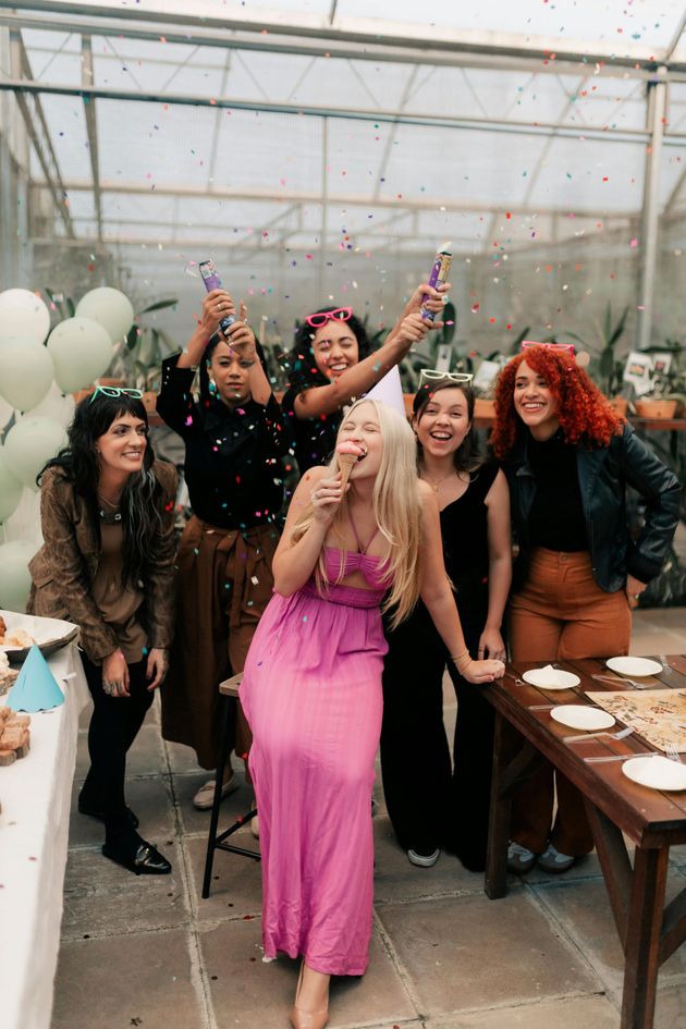 Group of six women celebrating, confetti exploding in the air. One woman in pink dress laughs; others smile, some holding party poppers.