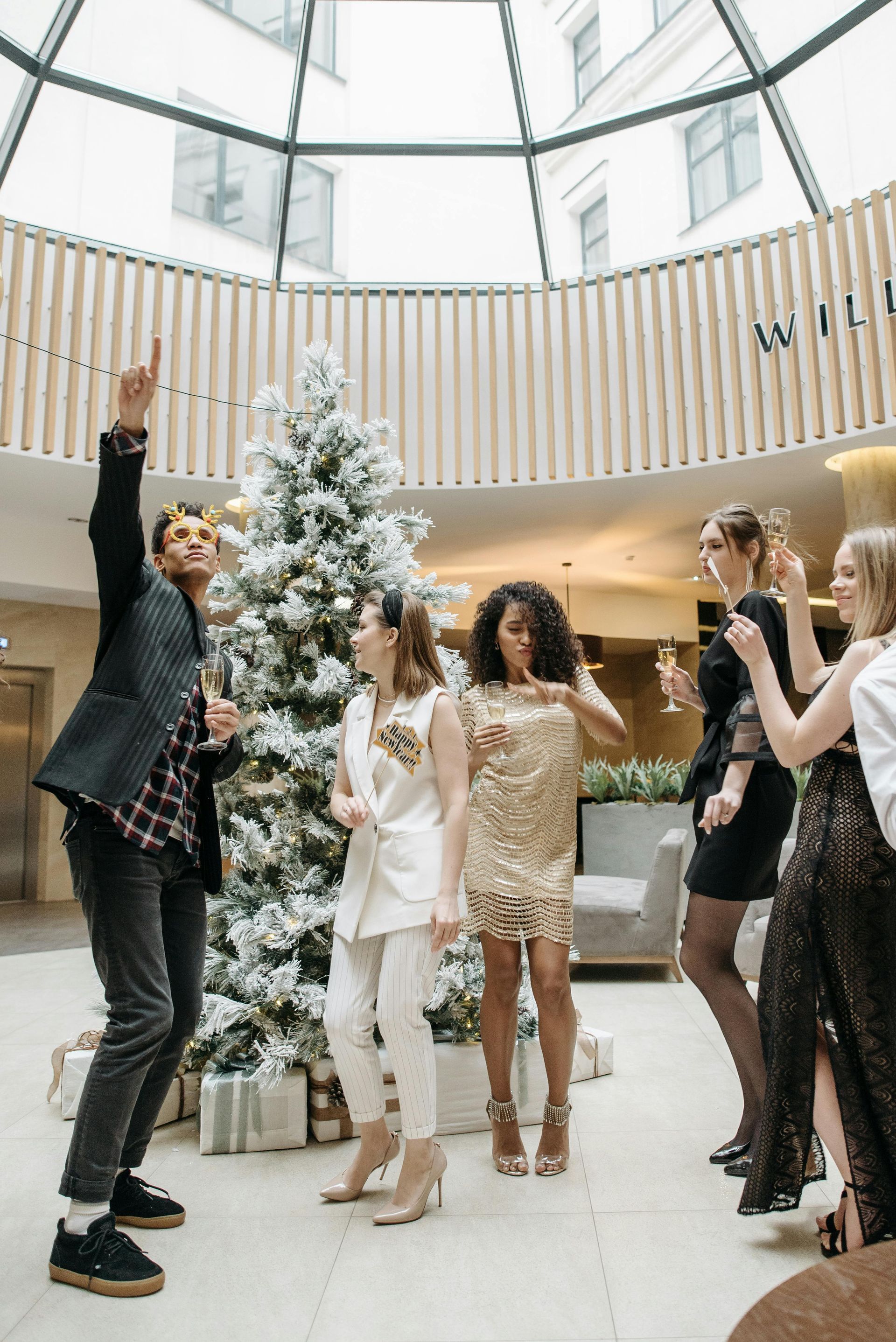 Group of people celebrating near a Christmas tree in a bright lobby, with a man raising an arm, smiling.
