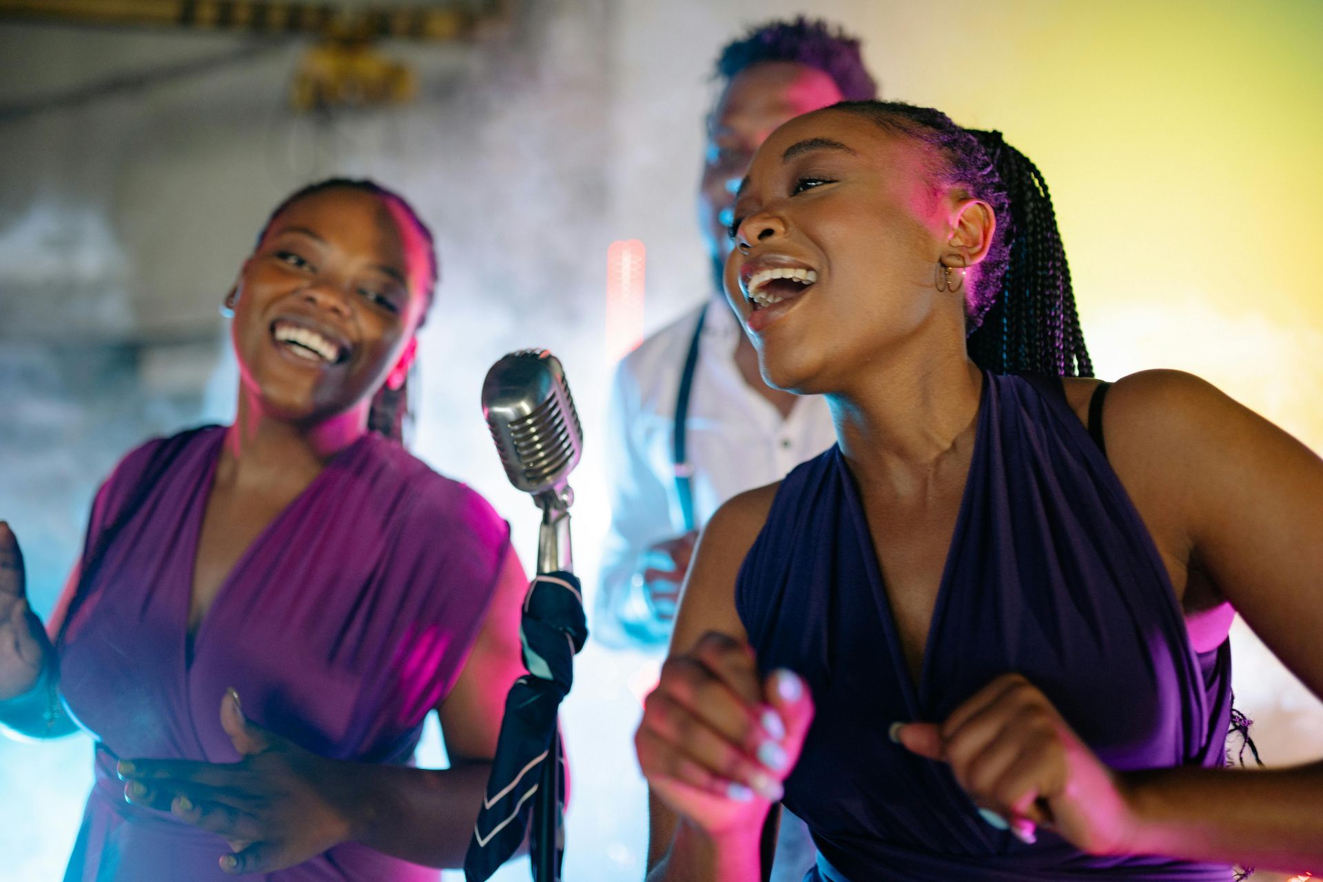 Three people singing and dancing on stage. Two women are in purple dresses, one is using a microphone. The setting is dimly lit with colorful lights.