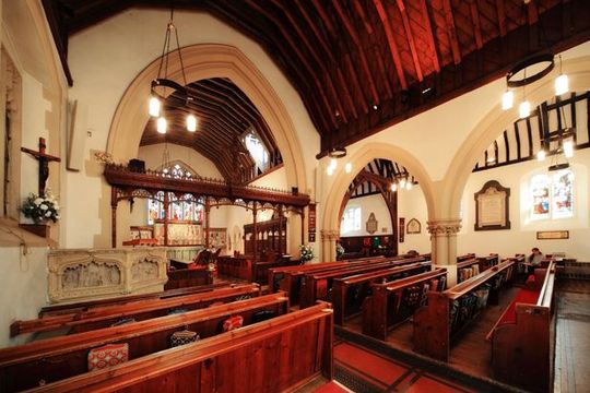 People in a church listen to two men at a table; large cross in window behind them.