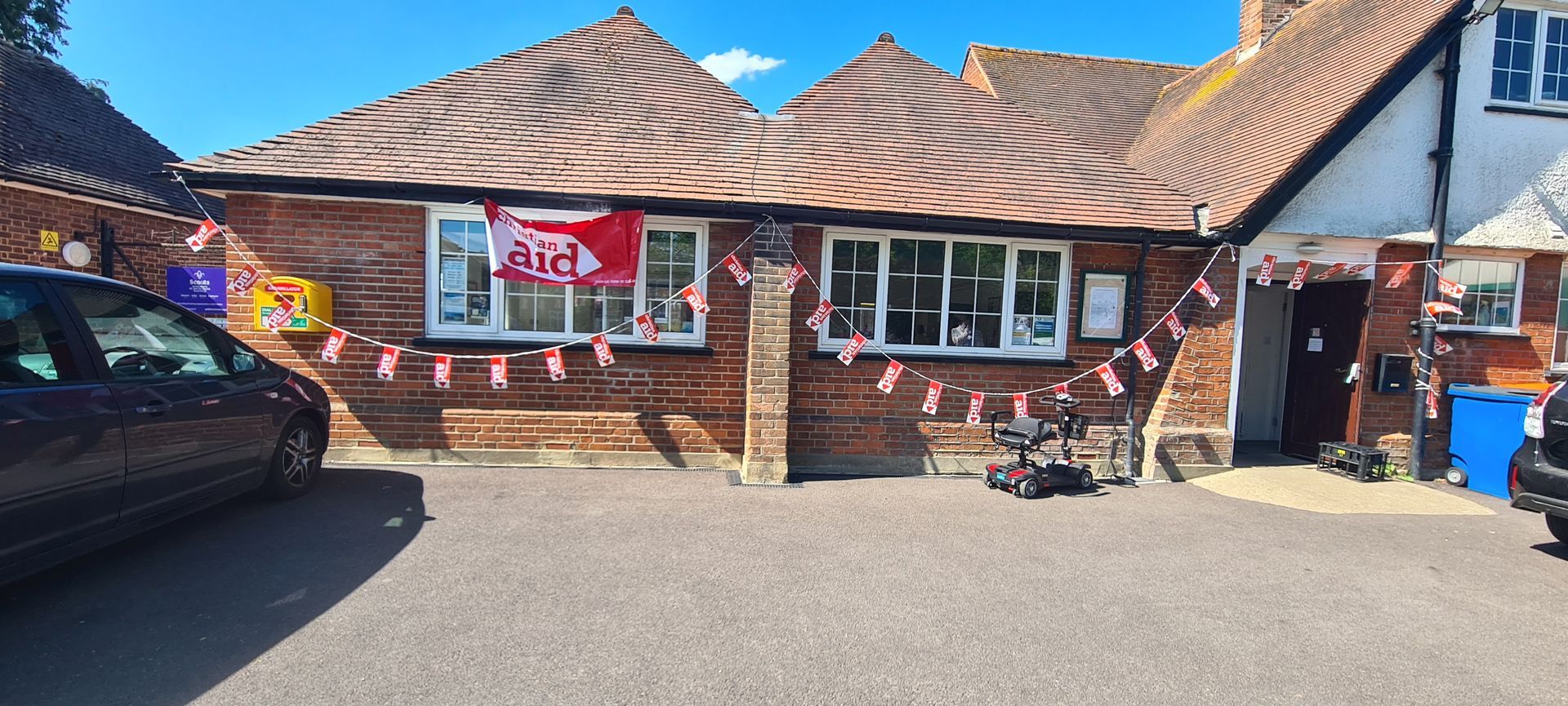 Exterior view of a brick building with a tiled roof. Bunting and a banner are displayed. A car is parked on the left.