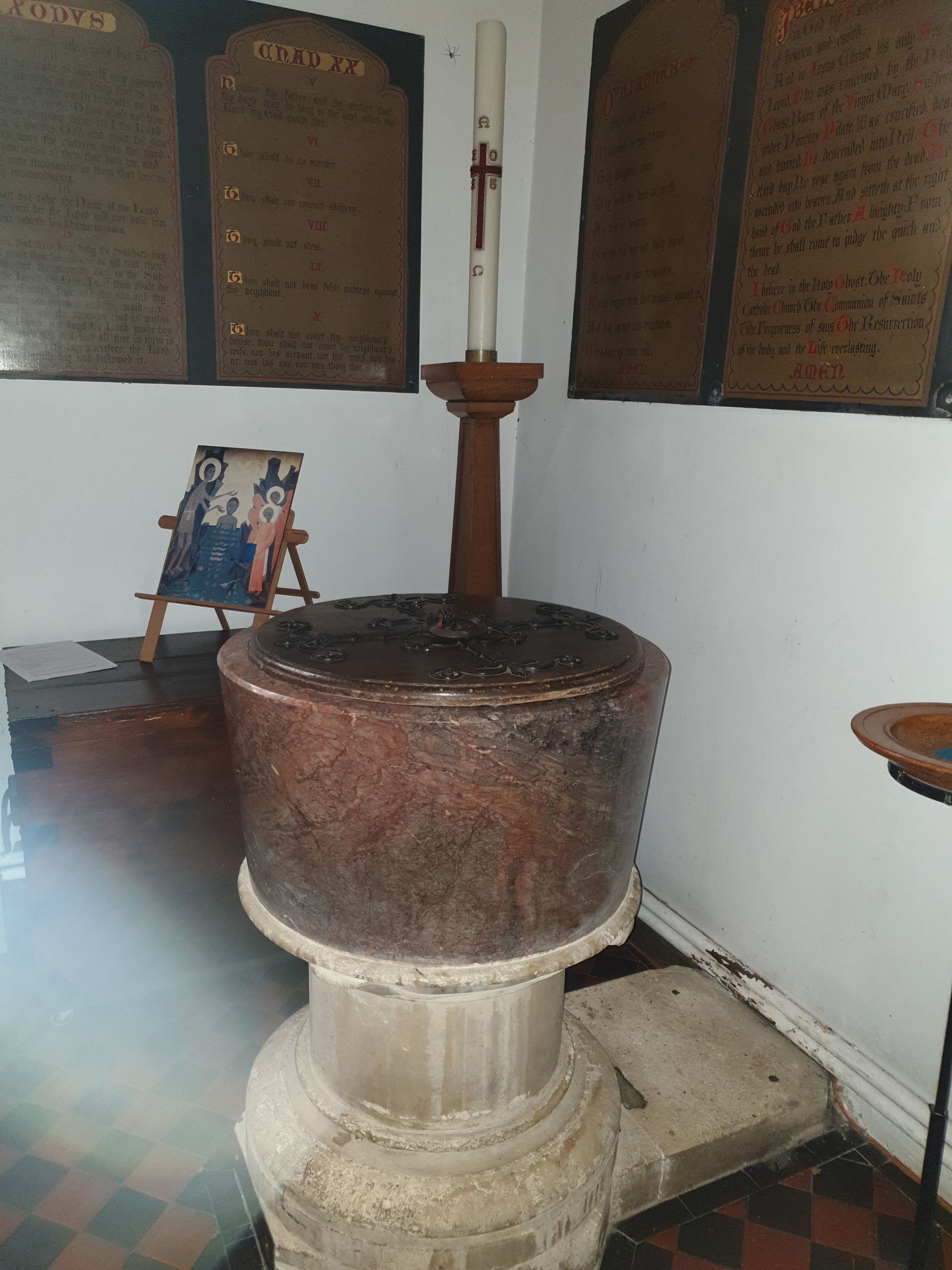 Baptismal font in a church, topped with a candle. Memorial plaques on the wall.
