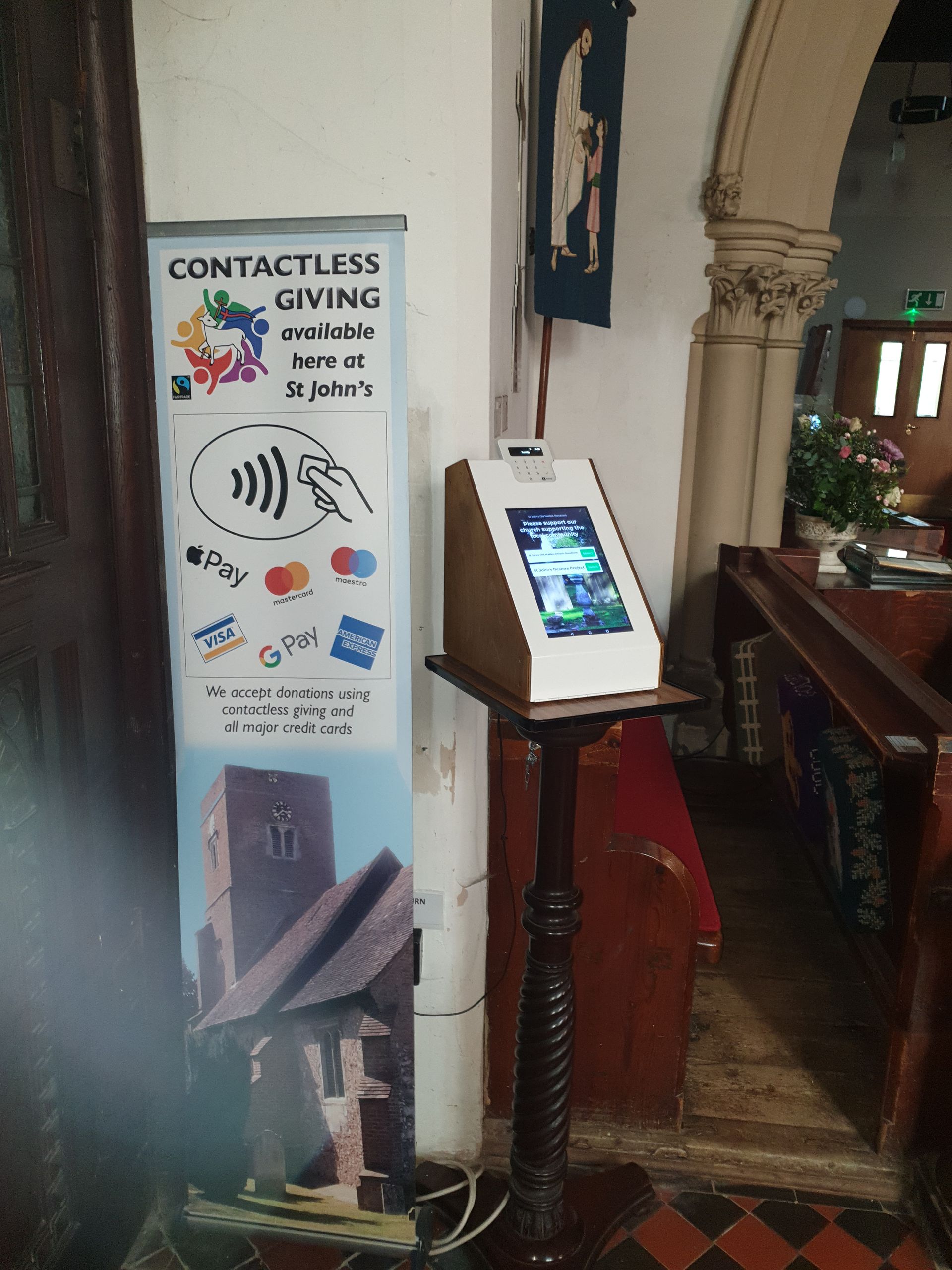 A contactless giving kiosk in a church next to a promotional banner and stained glass.