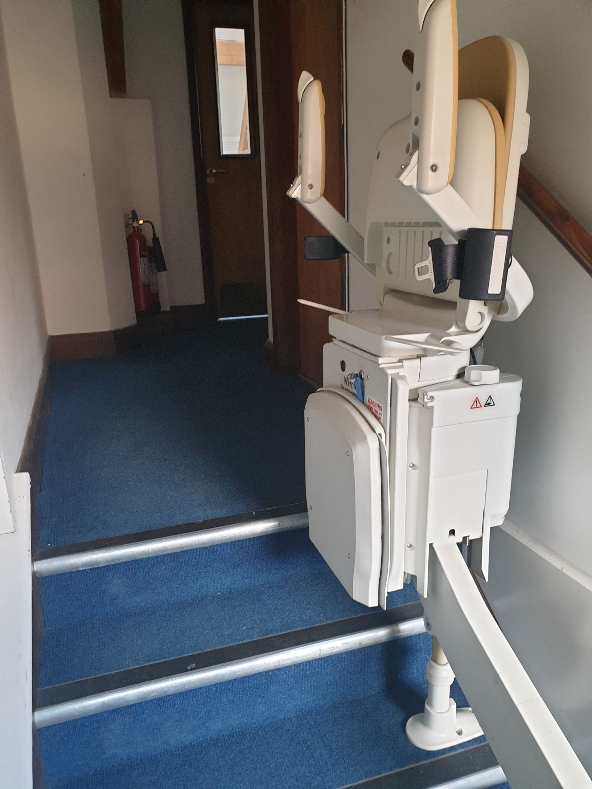 Stairlift on blue carpeted stairs. White mechanism with seat folded against wall. Wooden door and hallway in background.