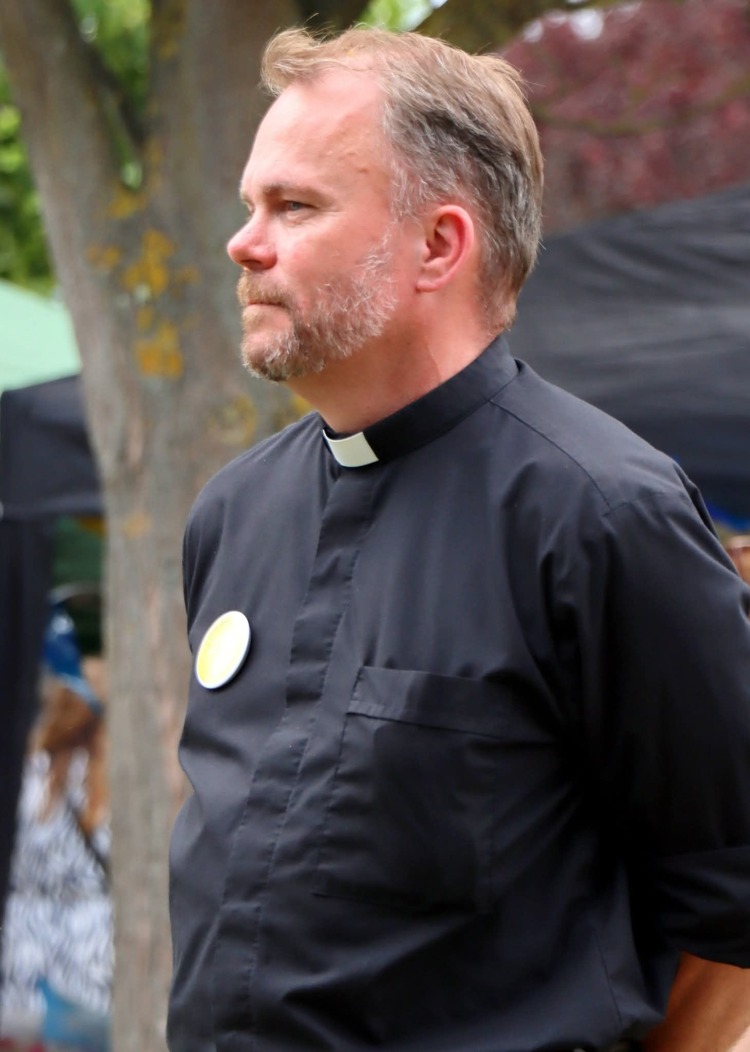 Man in black clerical shirt with white collar, small badge. Standing outdoors, looking right.
