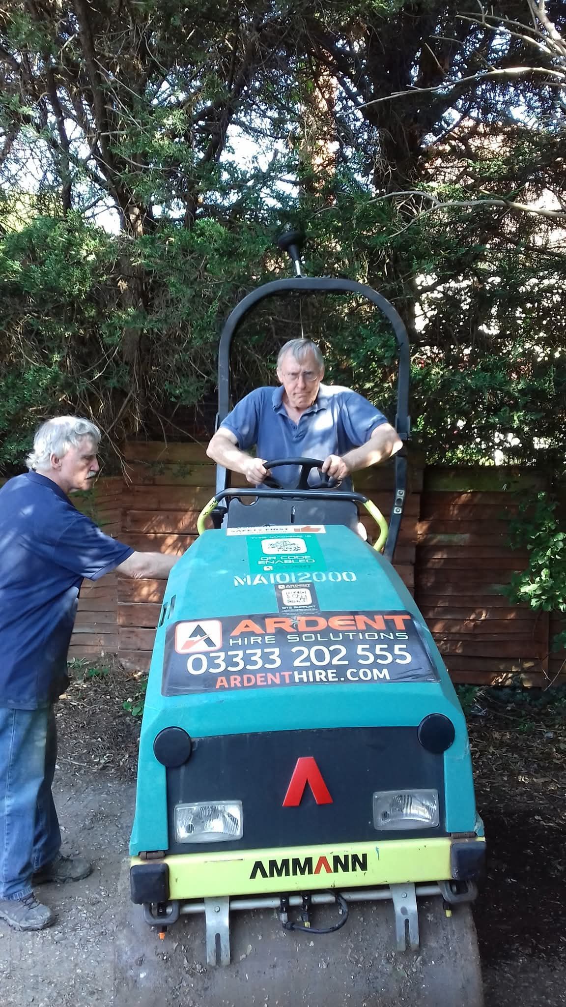 Man operates a small road roller while another man watches. They are in a yard.