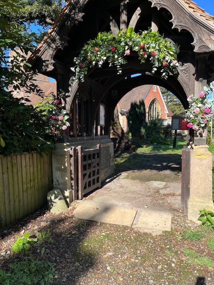 Wooden church entrance archway, decorated with flowers, leading to a path and church building.
