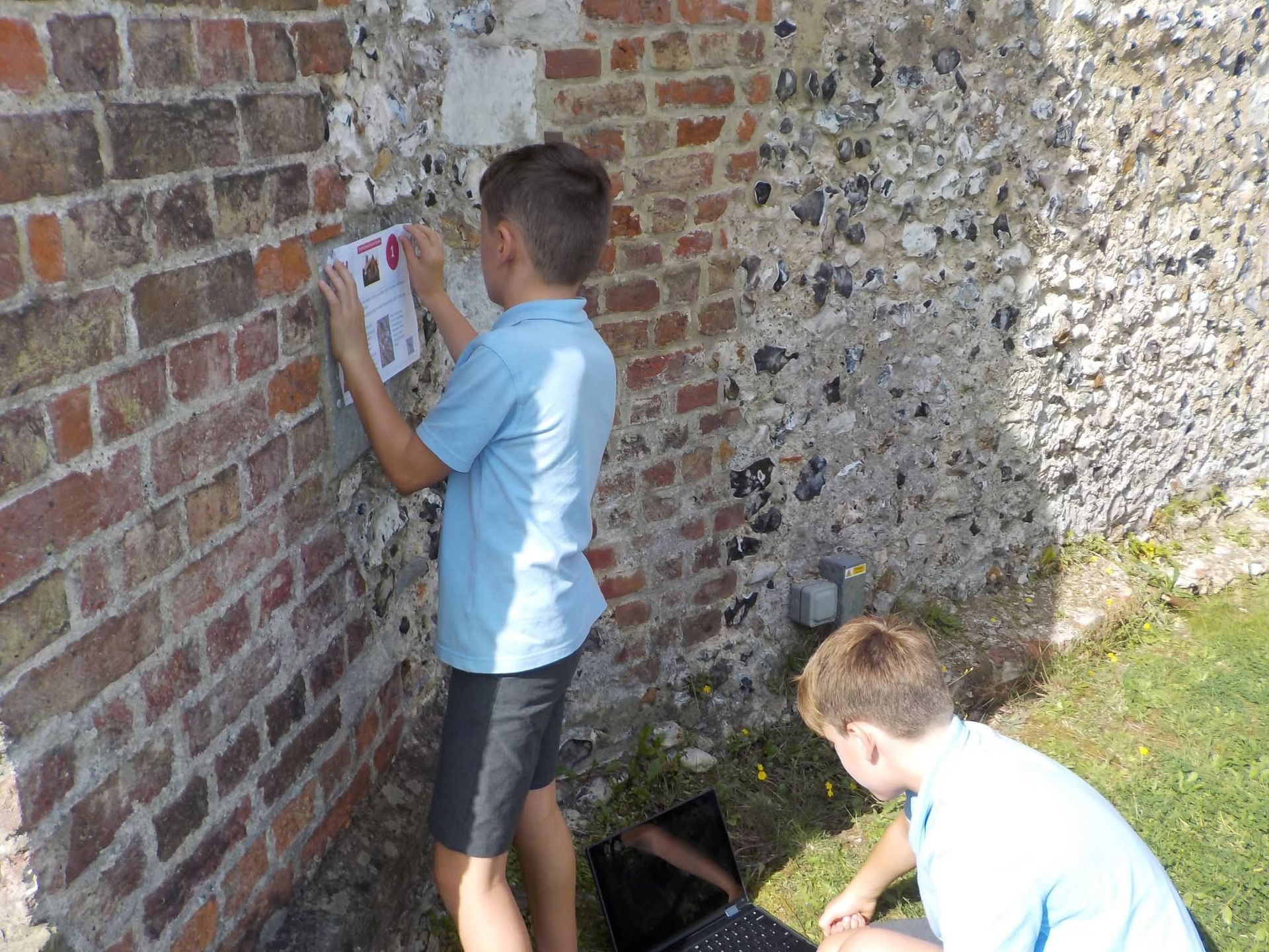 Two boys by a brick and stone wall, one placing a paper on the wall, the other looking at something. Sunny outdoors.