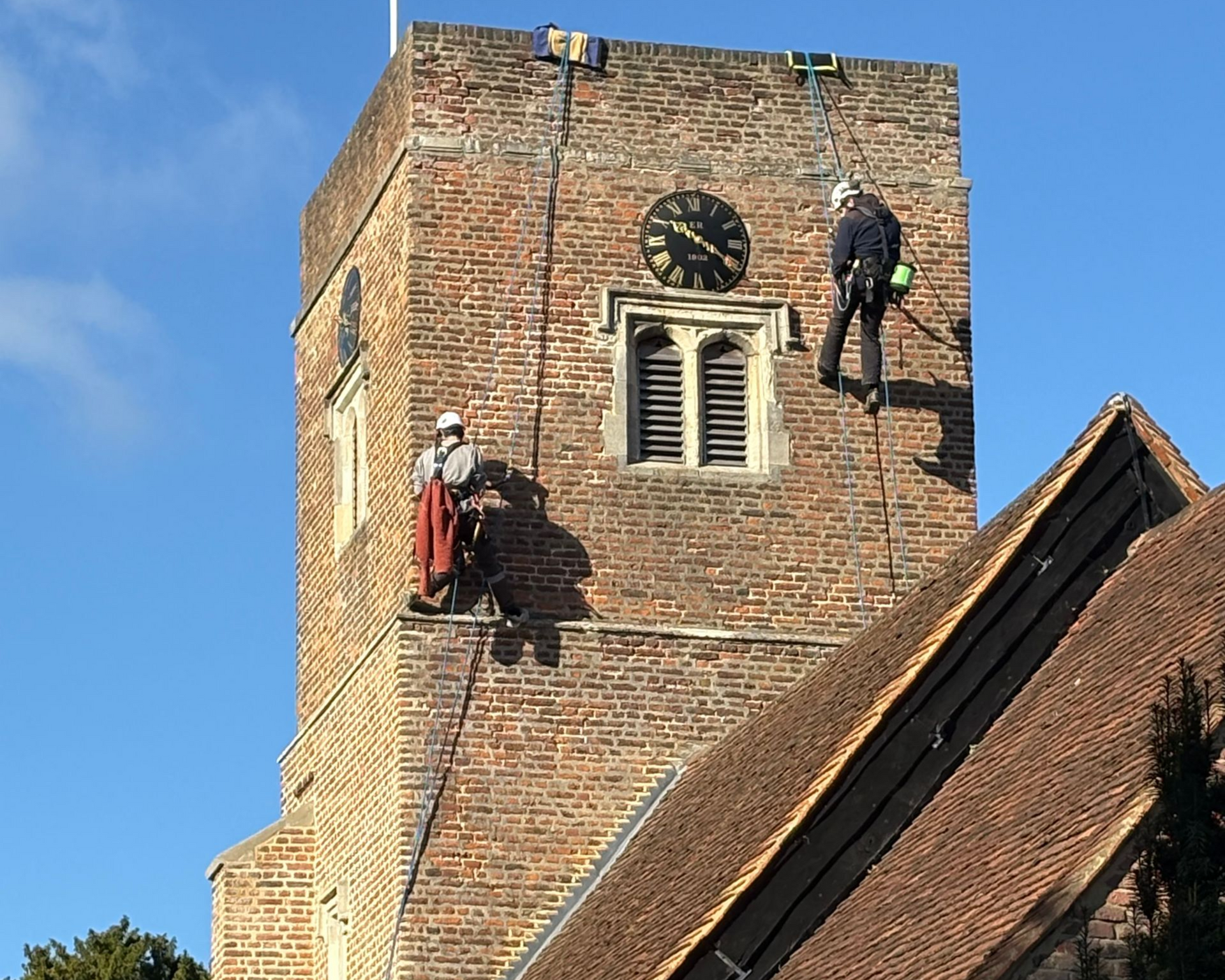 Two people on ropes inspecting a brick church tower with clock and roof. Blue sky.