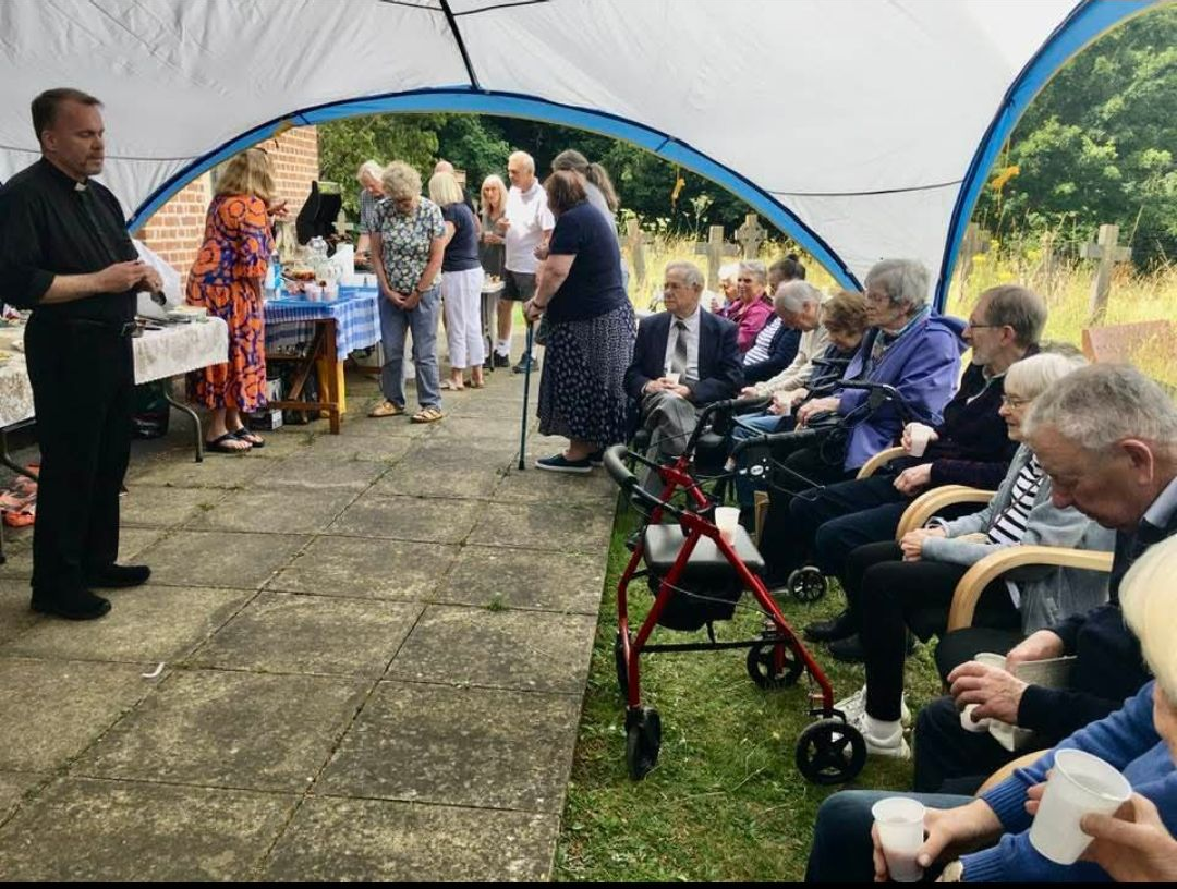Outdoor gathering under a tent, with a man in black speaking to a seated audience. Tables with food visible.