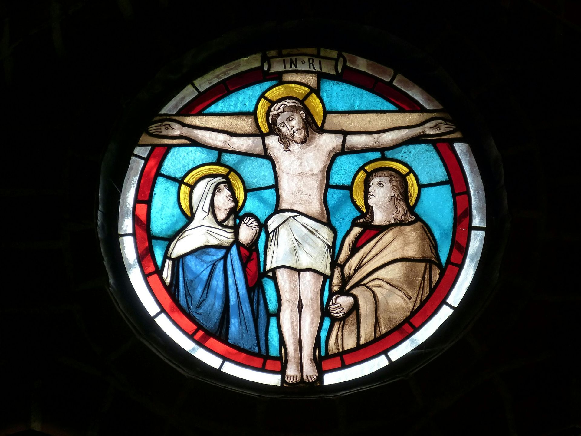 Stained glass depicting Jesus on the cross, flanked by two figures, set in a round window, blue and red hues.