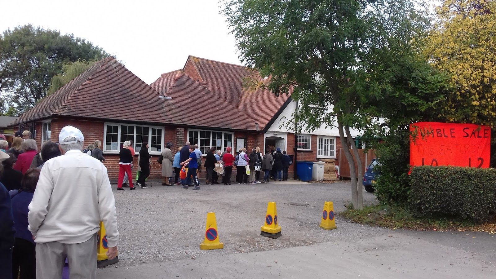 People waiting in line at a building with a "Jumble Sale" sign. Cones mark the entrance.