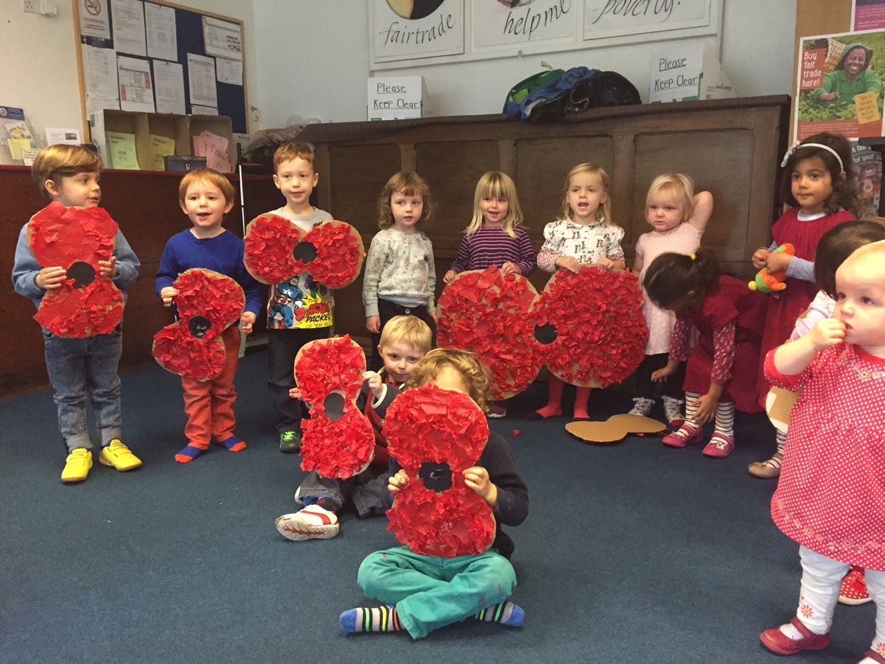 Children in a classroom holding large paper poppies. Some are standing, others kneeling, all smiling.