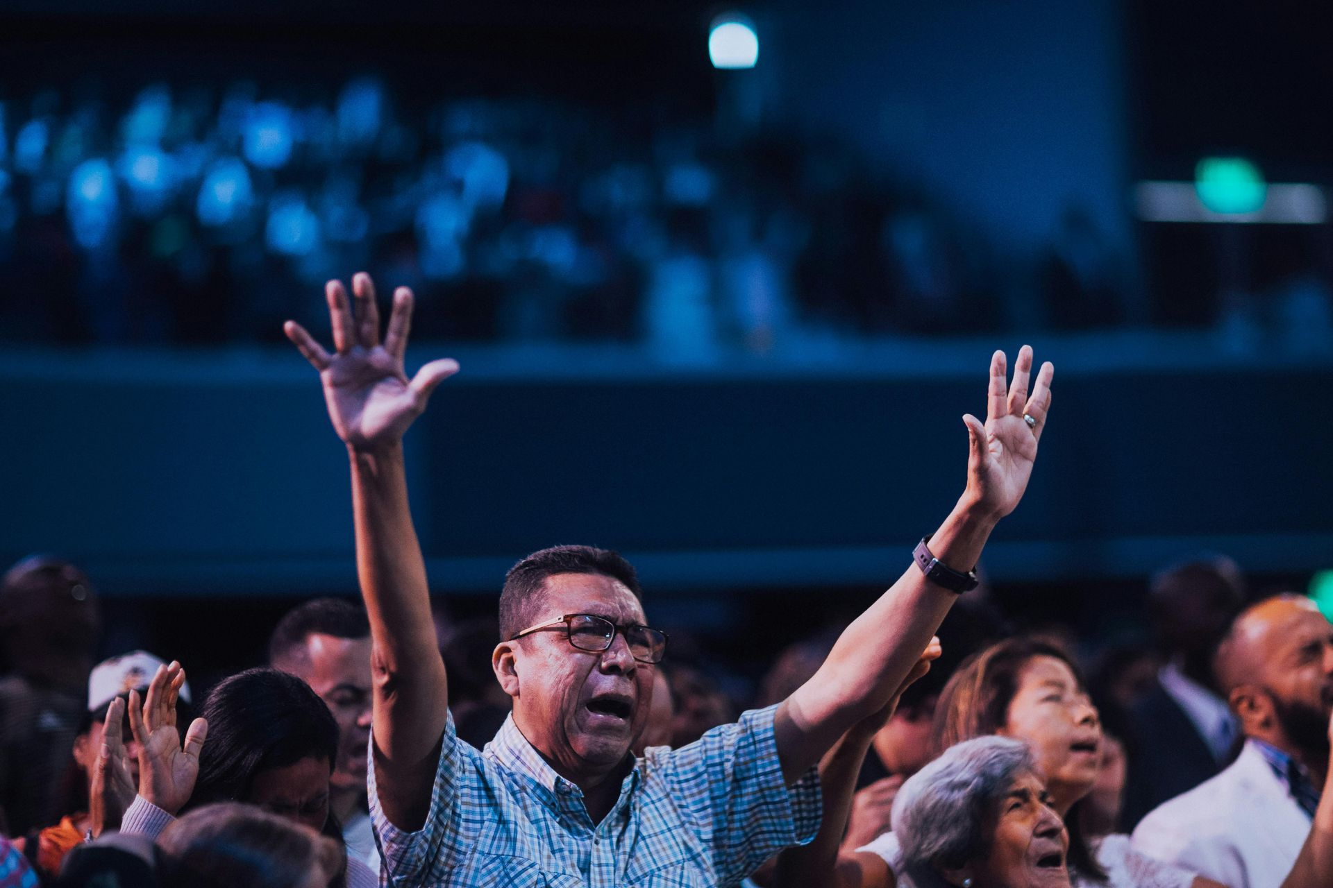 Person with arms raised in worship, surrounded by a crowd in a large auditorium, blue lighting.