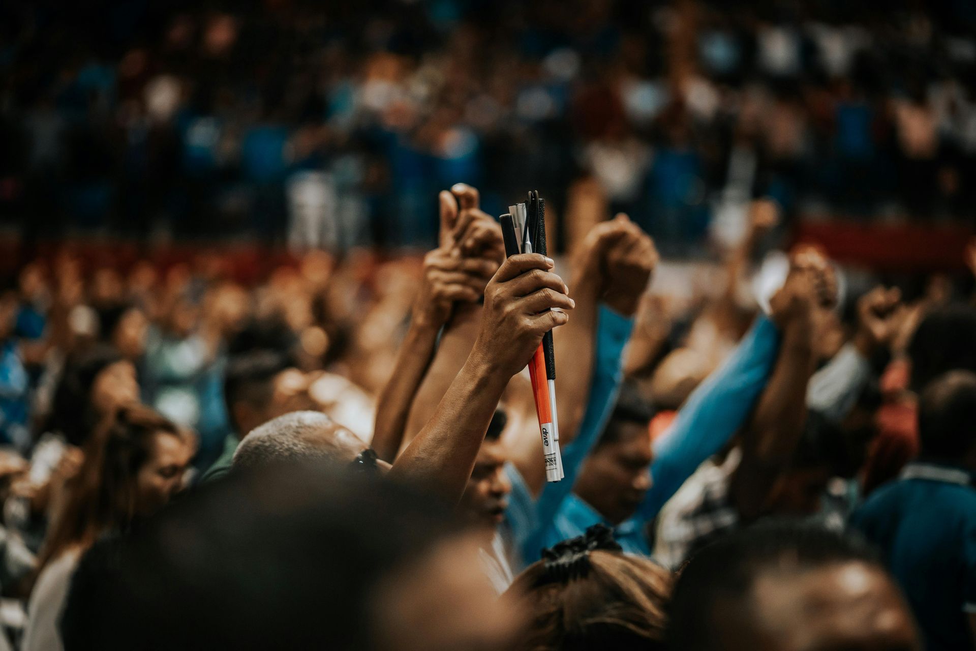 Hands raised, holding objects, crowd in background at an event with blue and red tones.