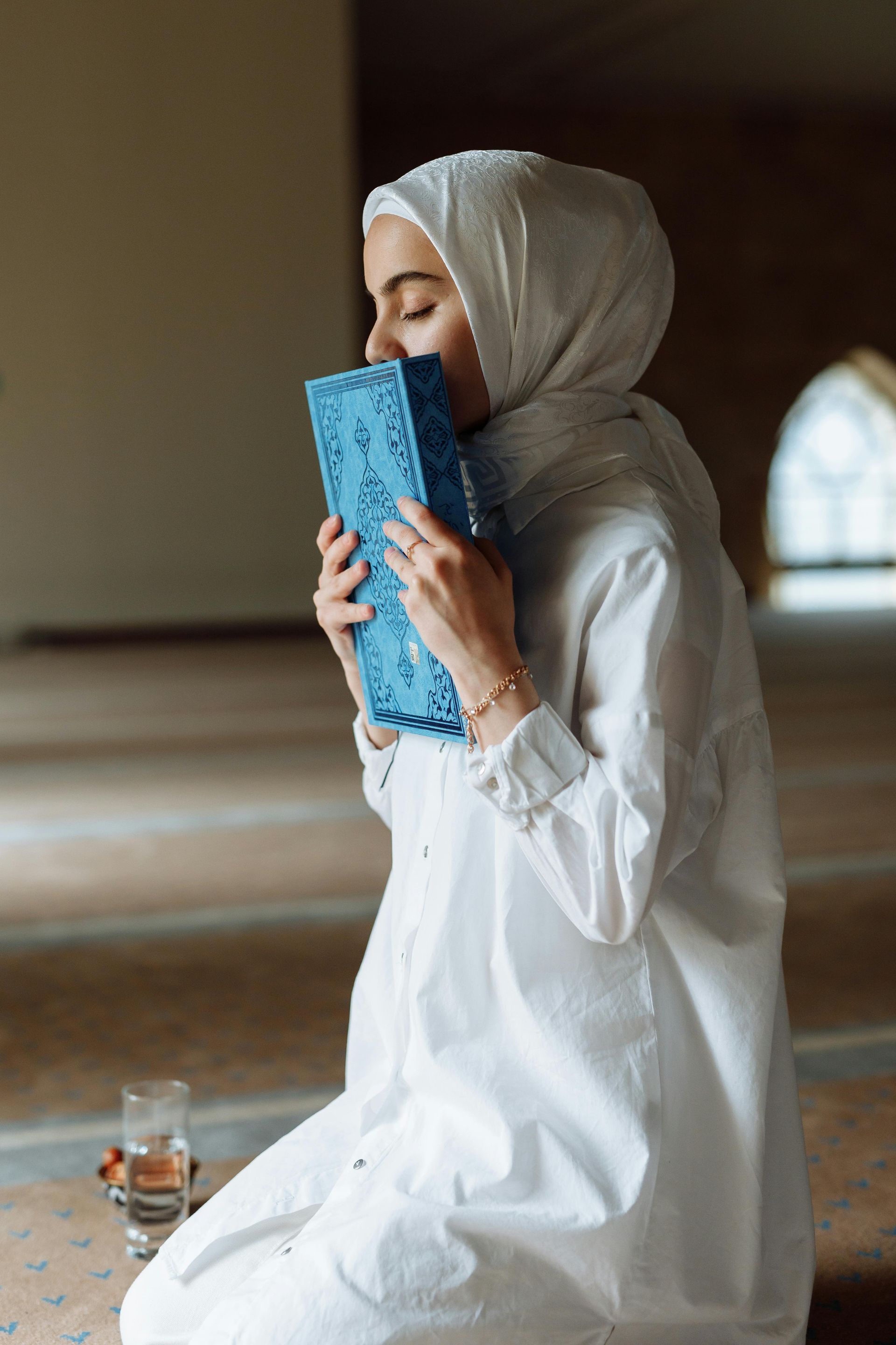 Woman in white clothing and hijab kneels, kissing a blue book, indoors near a glass of water.