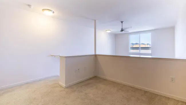Empty loft interior with white walls, tan carpet, and a ceiling fan near a window.