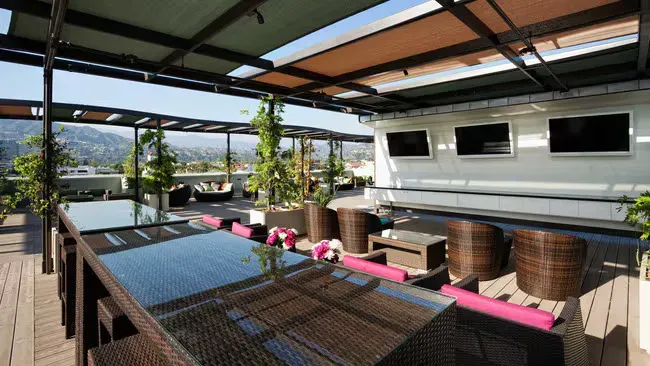 Rooftop patio with a long table, seating, TVs, and plants. Overlooking a city and mountains on a sunny day.