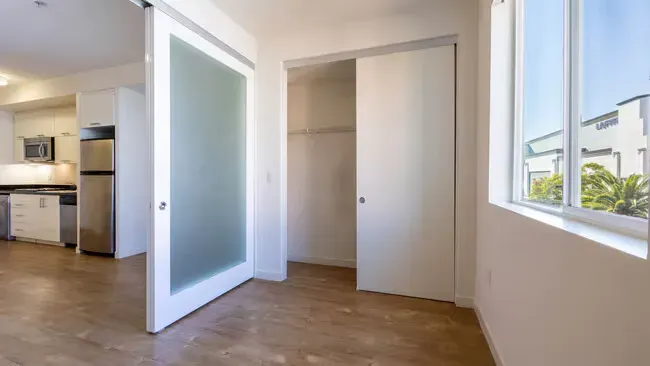 Empty closet with sliding doors, visible kitchen and window to the right.