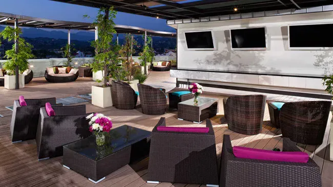 Rooftop bar with wicker furniture, flowers, and city views at dusk.