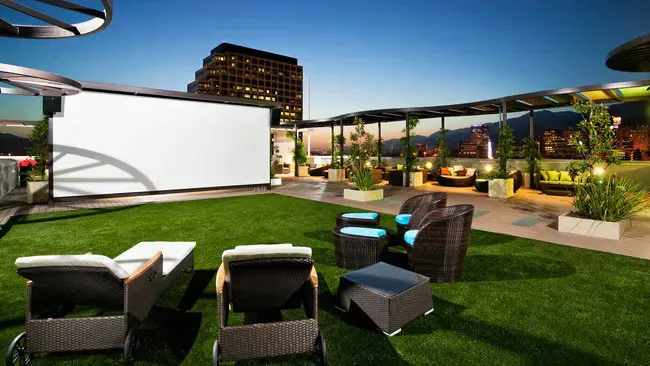 Rooftop outdoor movie theater with artificial grass, seating, screen, and city skyline at dusk.
