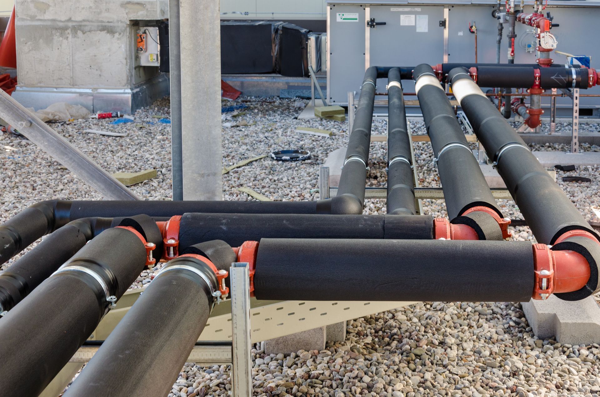Insulated pipes with red fittings, supported by metal framing, outdoors against a building.