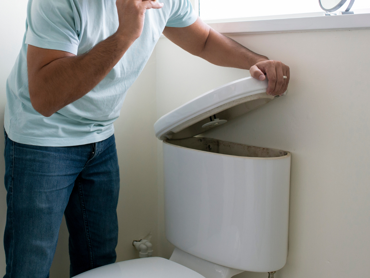 Man in jeans and a light blue shirt looking into an open toilet tank in a bathroom. Man in jeans and a light blue shirt looking into an open toilet tank in a bathroom.