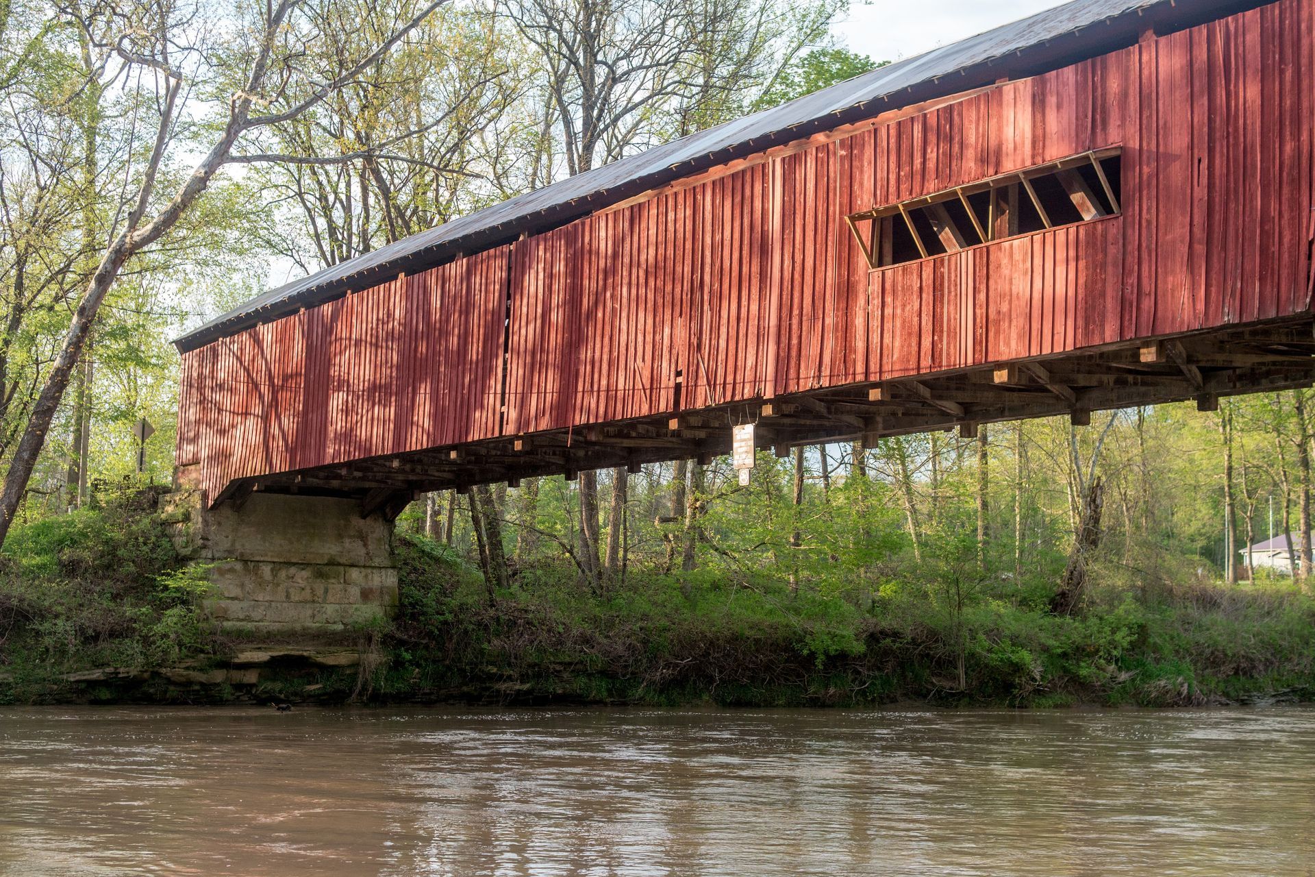 Red covered bridge over a river, with trees lining the banks and sunlight casting shadows.