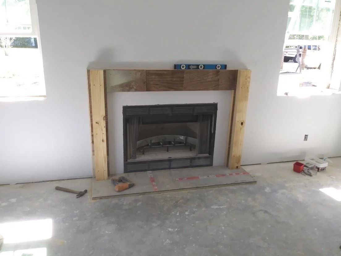 Fireplace under construction, framed with wood, in a room with two windows and unfinished walls.