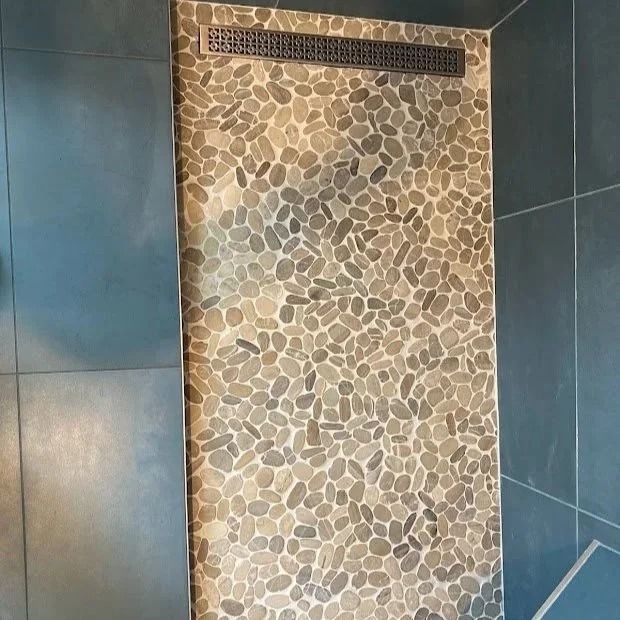 Shower with pebble tile floor and linear drain, surrounded by blue-gray tiled walls.