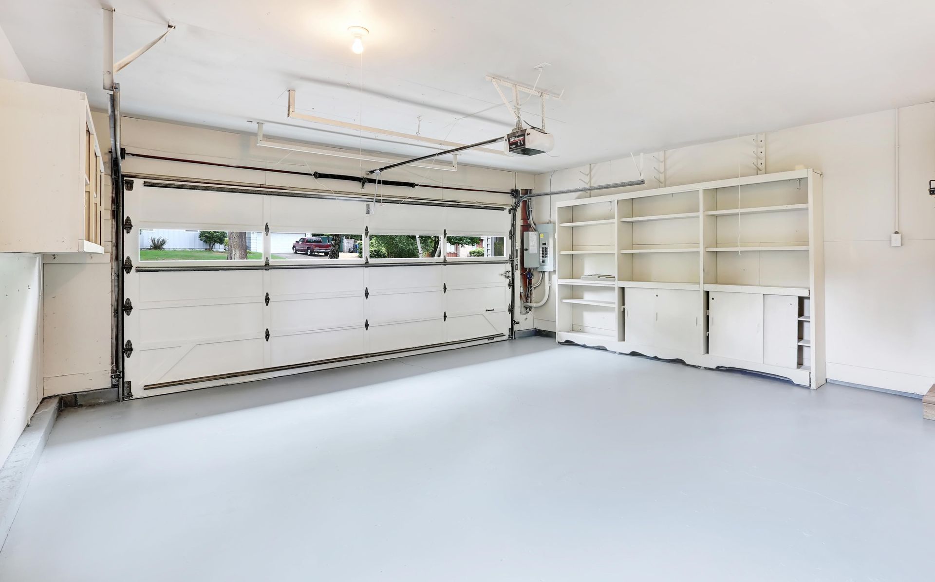 Empty white garage with gray floor, white cabinets, and open garage door.
