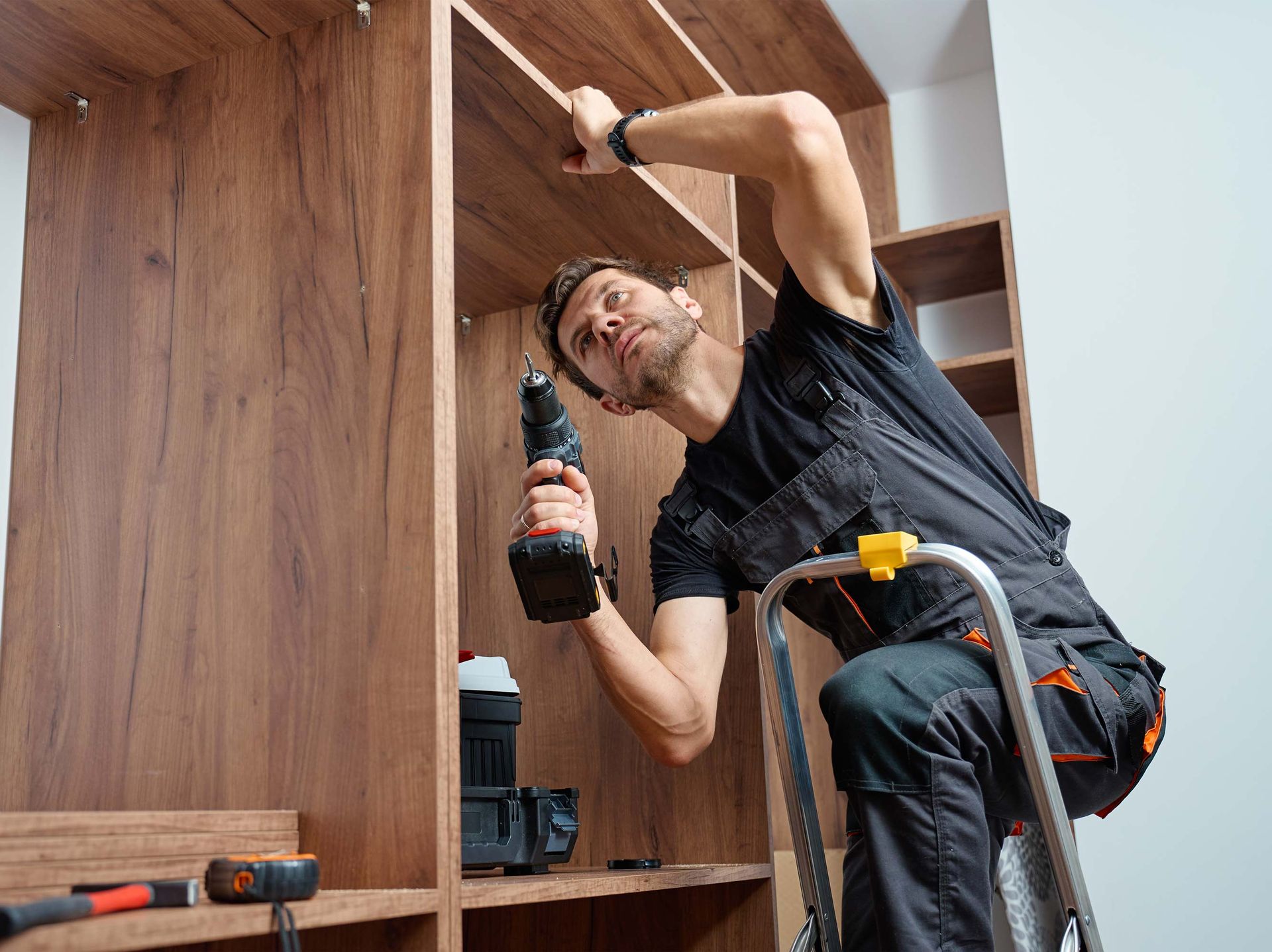 A person on a step ladder using a drill to assemble a wooden cabinet.