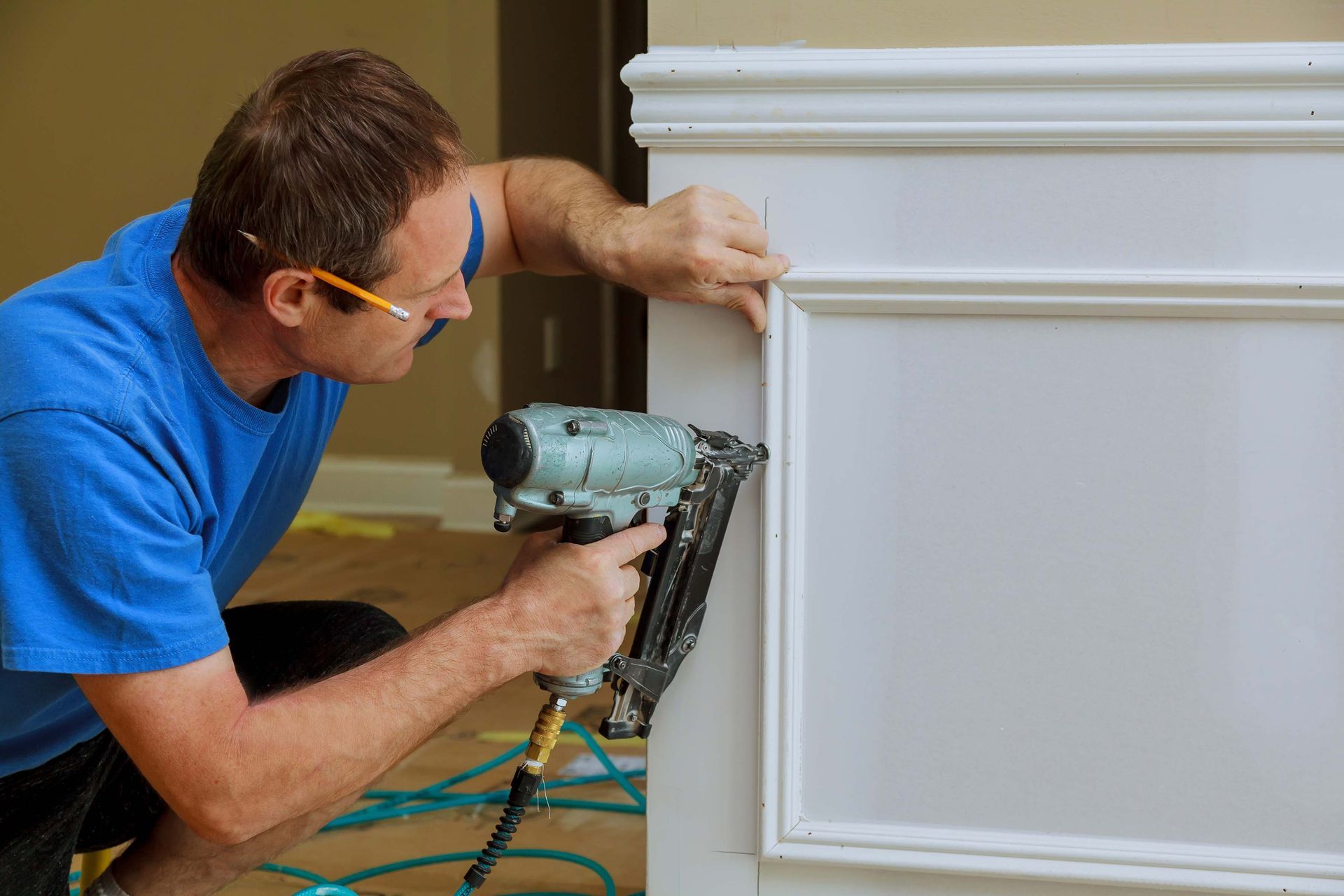 Man using a nail gun to install trim on a white wall in a room.