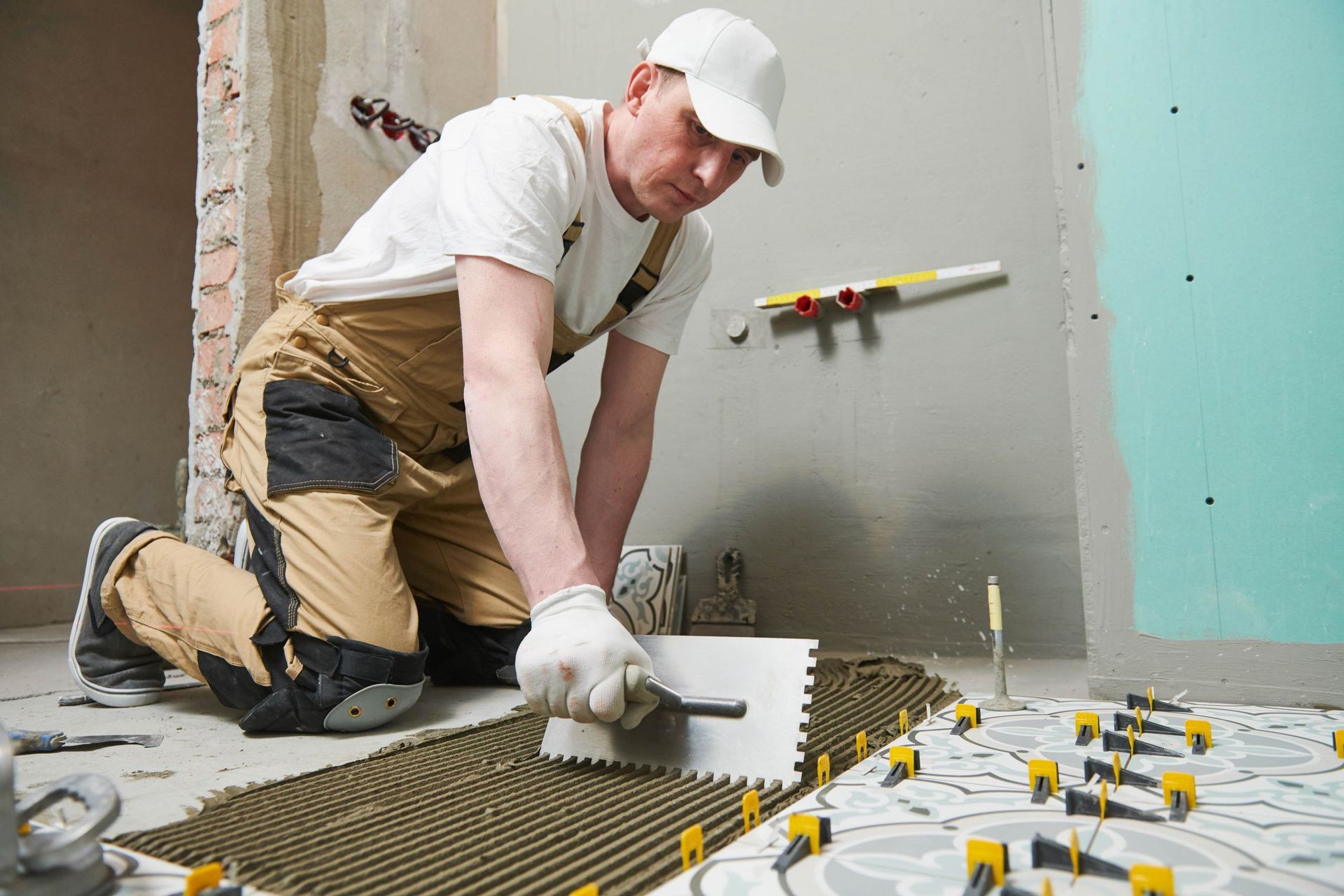 Man tiling a floor, applying adhesive with a notched trowel. The setting is a construction site.