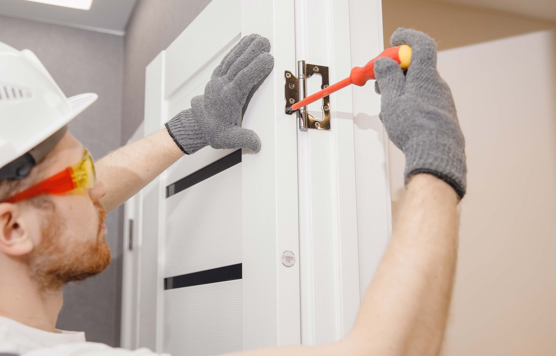 Person in gloves and hard hat screws door hinge with screwdriver.