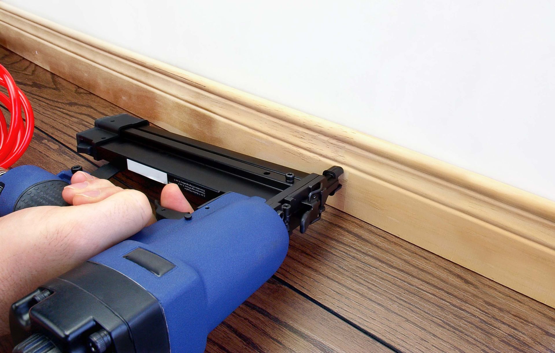 Person using a blue nail gun to install light-colored wood baseboard trim on a white wall above a wood floor.