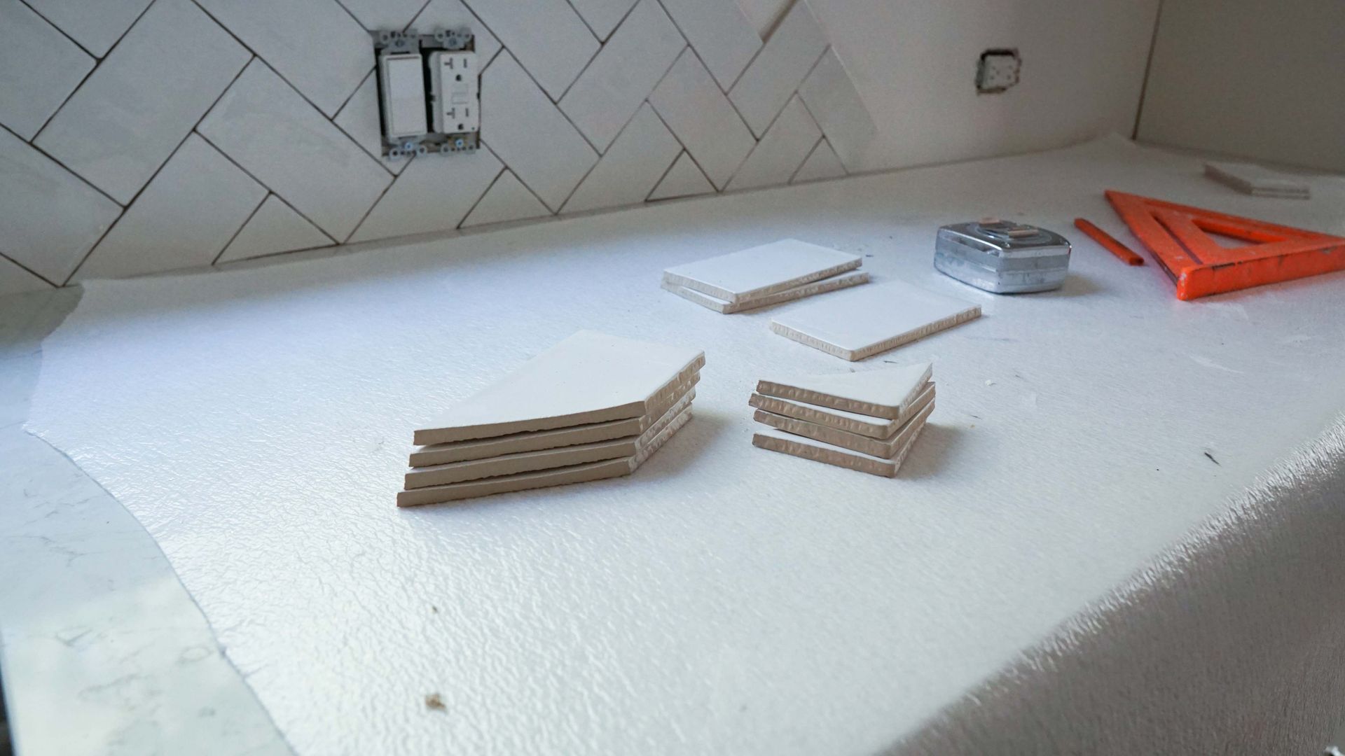Countertop with white herringbone tile backsplash under construction; tools and tiles are present.