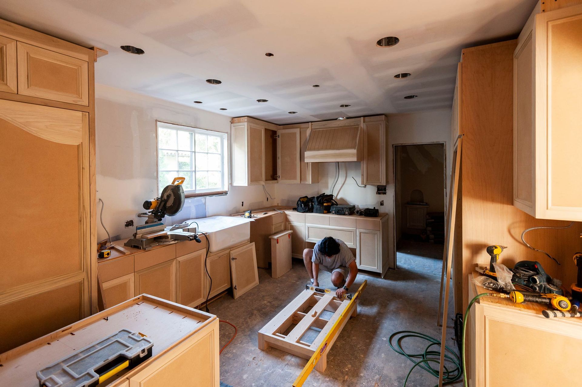 Kitchen under renovation with cabinets, tools, and a person working on a wood frame.
