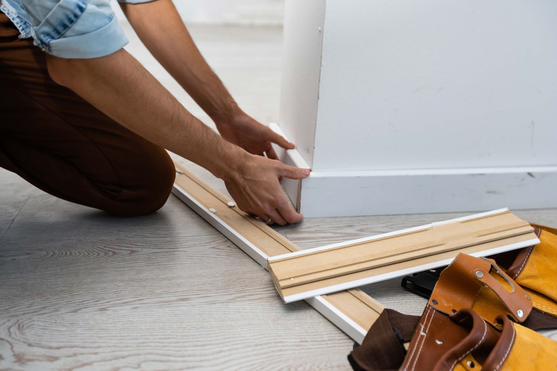 Person installing baseboard molding on a white wall, with a tool belt visible.