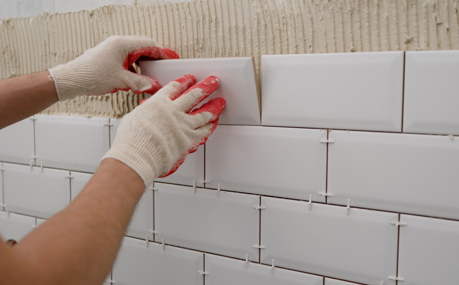 Person placing white subway tiles on a wall, using spacers and wearing gloves.